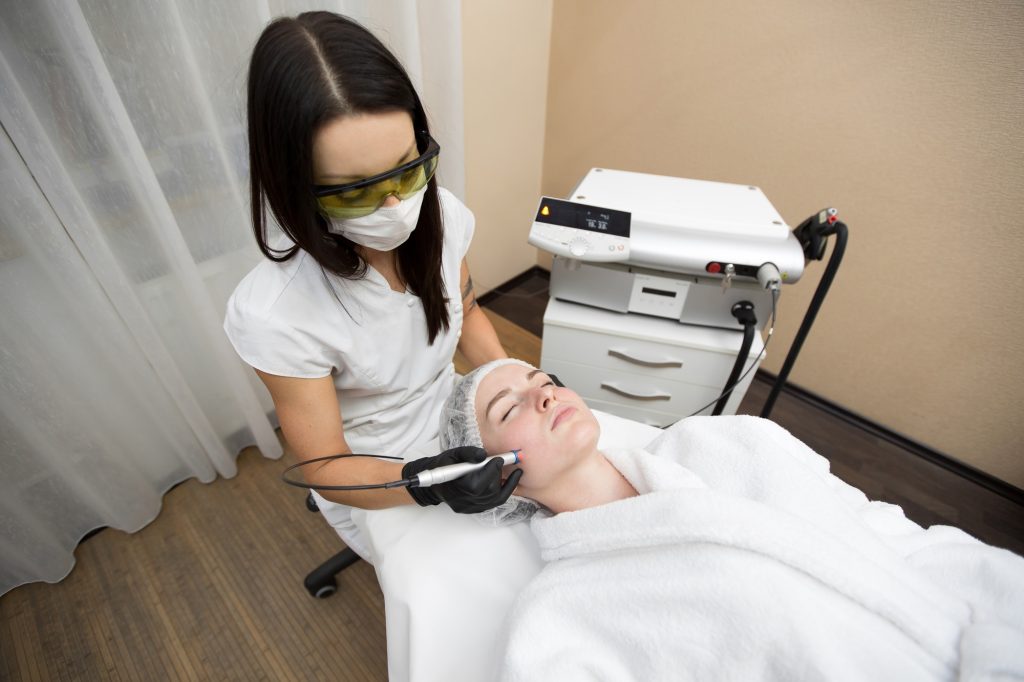 Cosmetologist in protective glasses conducts laser treatment of the skin for a young patient